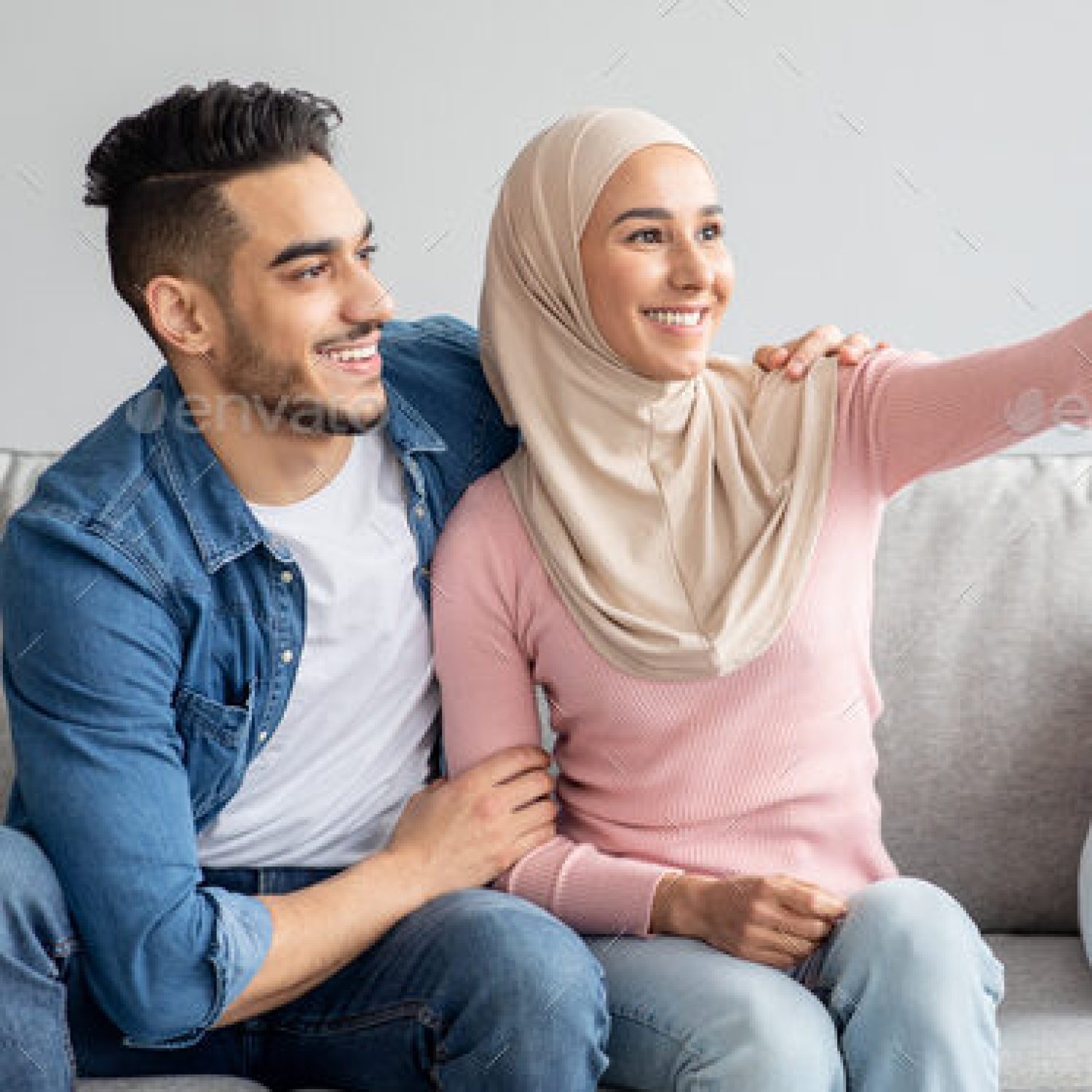 Handsome middle-eastern man and woman in hijab smiling at smartphone camera, copy space. Beautiful arab couple embracing and taking selfie on mobile phone for social media while resting at home